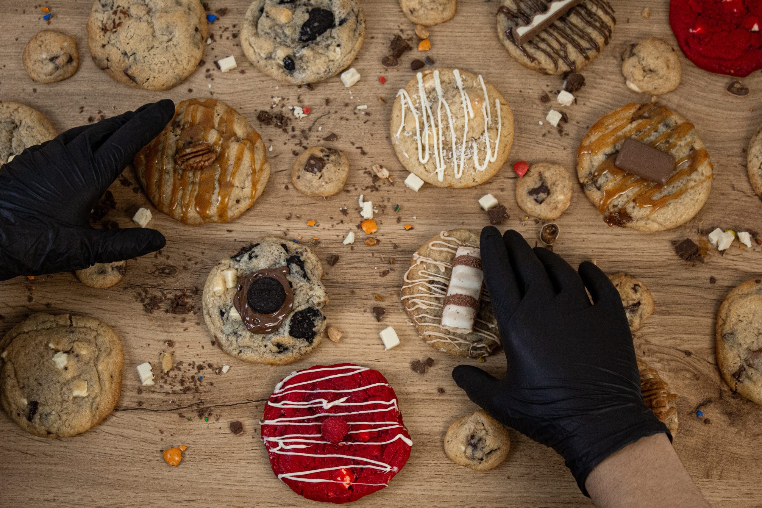 Deux mains gantées de noir au-dessus d’un plan de travail en bois couvert de cookies : pécan, chocolat blanc, glaçage rouge.